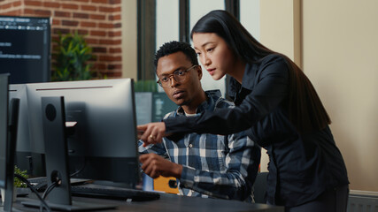 Two software developers coming at desk and sitting down holding laptop with coding interface and pointing at computer screen. Programmers team discussing algorithms looking at monitors with code.