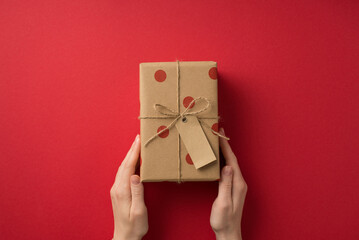 First person top view photo of valentine's day decorations woman's hands giving craft paper giftbox with polka dot pattern and twine bow on isolated red background with copyspace