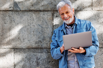 A happy senior student is standing against the brick wall and typing on a laptop.