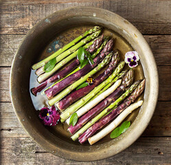 Set of raw white, green, purple asparagus in copper basin with violet and mint. Old wooden rustic background. Organic healthy eating concept. Overhead.