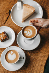 Woman is holding a cup of coffee near cheesecake and brownie placed on a wooden table