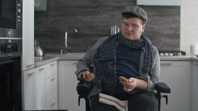 Locked-down of Caucasian man in wheelchair wearing eyeglasses and peaked cap, taking cookies from baking sheet in oven at daytime in his modern kitchen at home and leaving frame