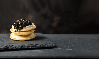 A stack of blini pancakes with black caviar and sour cream on a slate, copy space