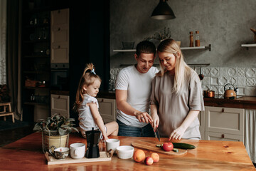 A happy family with one child having breakfast laughing sitting at the dining table in a loft-style kitchen in a cozy house. Pregnant mom dad and little daughter cook together. Selective focus