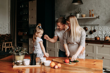 A happy family with one child having breakfast laughing sitting at the dining table in a loft-style kitchen in a cozy house. Pregnant mom dad and little daughter cook together. Selective focus