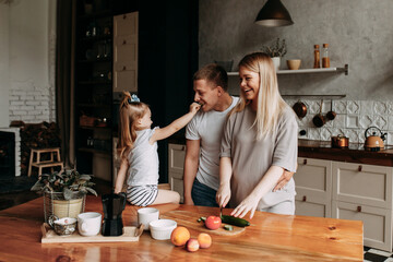 A happy family with one child having breakfast laughing sitting at the dining table in a loft-style kitchen in a cozy house. Pregnant mom dad and little daughter cook together. Selective focus