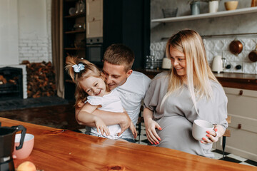A happy family with one child having breakfast laughing sitting at the dining table in a loft-style kitchen in a cozy house. Pregnant mom dad and little daughter cook together. Selective focus