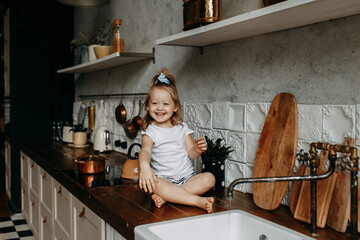 A cheerful little girl in pajamas is sitting on the kitchen cabinet playing with water from the...
