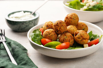 Salad bowl with chickpea falafel, fresh green spinach, cherry tomatoes, croutons and sour cream dressing on a grey stone surface
