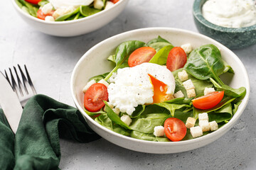 Salad bowl with fresh green spinach, cherry tomatoes, croutons, egg benedict and sour cream dressing on a grey stone surface