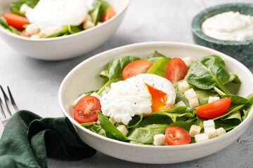 Salad bowl with fresh green spinach, cherry tomatoes, croutons, egg benedict and sour cream dressing on a grey stone surface
