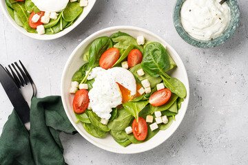 Salad bowl with fresh green spinach, cherry tomatoes, croutons, egg benedict and sour cream dressing on a grey stone surface