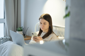Young woman using laptop computer and surfing social media on smartphone, sitting on bed at home