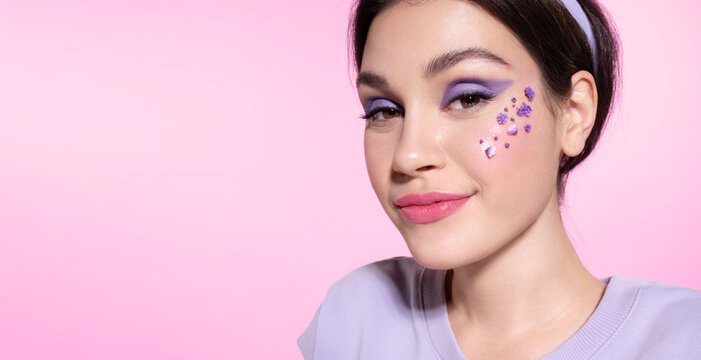 Modern young teen girl with lilac eyeshadow and flowers on healthy smooth cheek skin. Cool woman with lavender make up smiling on pink background. Skincare, make-up cosmetics advertising for youth.
