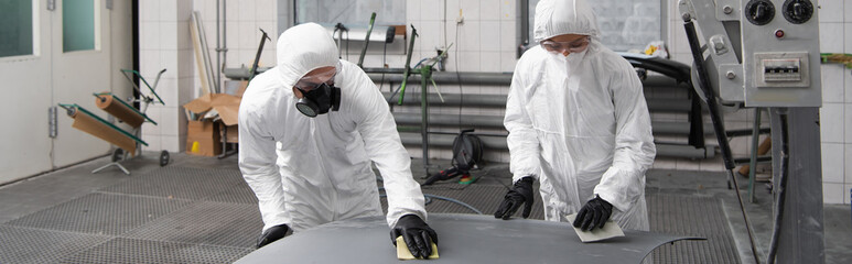 Mechanics in protective suits working with sandpaper and car hood in garage, banner.