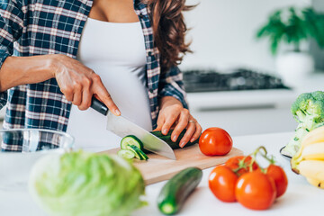 Unrecognisable pregnan woman cook salad cutting cucumber on wooden cutting board putting fresh vegetables and fruit on table in kitchen. Pregnancy, maternity, healthy lifestyle concept.