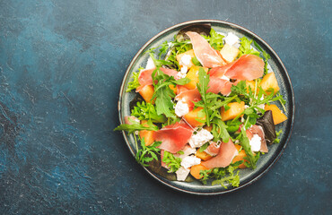 Summer salad with cantaloupe melon, prosciutto, white cheese and arugula on blue table background, top view, copy space