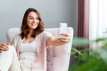Beautiful tender young smiling caucasian 30s woman taking picture selfie using cell phone near window. Long haired brunette is photographing yourself on smartphone home interior. - obrazy, fototapety, plakaty