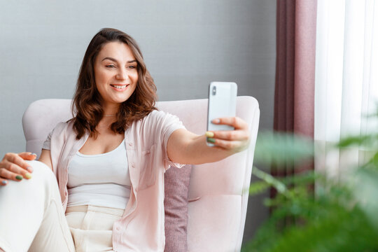 Beautiful tender young smiling caucasian 30s woman taking picture selfie using cell phone near window. Long haired brunette is photographing yourself on smartphone home interior.