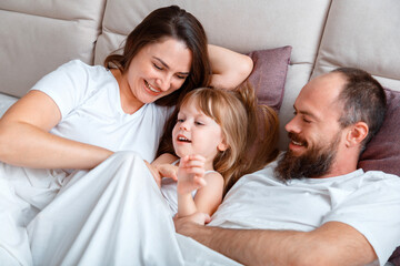 Young family play spend time together hugs on couch in morning. Mother Father and Daughter child have fun in Morning Routine at home in bedroom. 