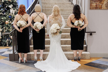 Bridesmaids and bride in a long white gown standing arm in arm with backs to the camera holding...