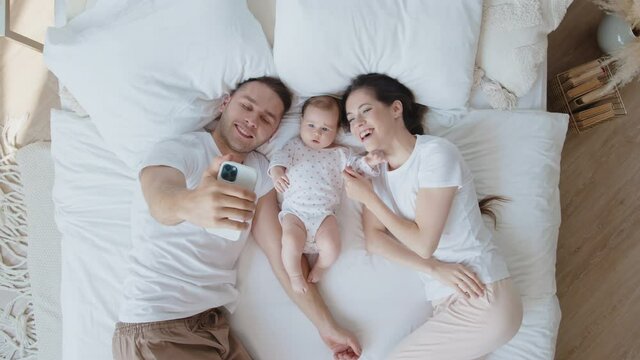 Young caucasian family with a small newborn baby are lying on white bed sheet, smiling, taking selfies or video call and waving hand at the smartphone camera. Concept of technology, new generation
