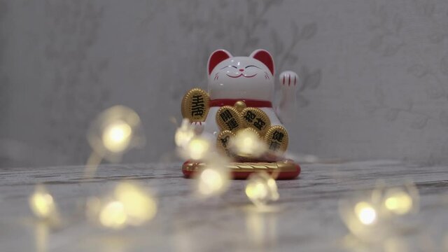 Chinese lucky cat and burning garland on blurred background. Maneki-neko ceramic cat symbolizing rich and wealth waves its left paw. 4K UHD video.