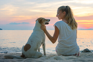 Young woman with dog sitting together on the beach and enjoying the sunset