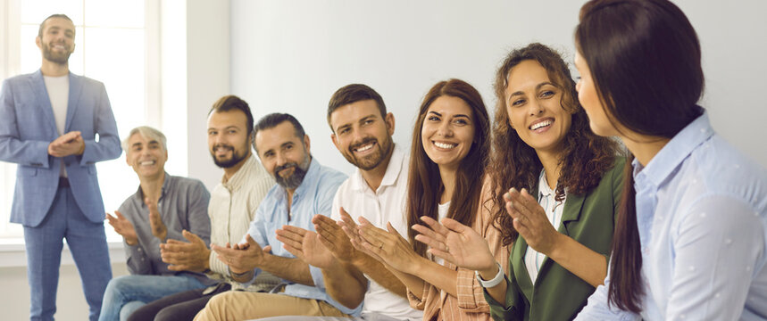 Positive smiling young and mature male and female team sitting in office applauding young woman for giving successful talk on business topic. Recognition and appreciation at work concept background