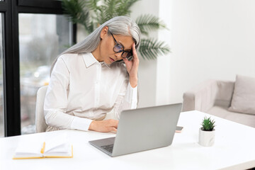 Tired mature female employee feel bored. Upset senior gray-haired woman sitting in front of the...