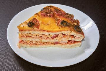 A piece of the three-layer homemade pizza with cheese, sausages, ham and olives. Close-up, selective focus.