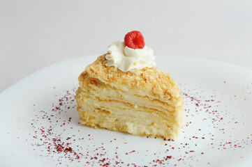 Slice of classic Napoleon cake decorated with cream and fresh raspberries. White plate, sprinkled with dried berries