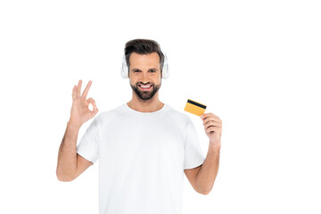 pleased man in headphones holding credit card and showing okay gesture isolated on white.