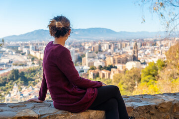Fototapeta na wymiar A young tourist sitting looking at the views of the city and the Cathedral of the Incarnation of Malaga from the Castillo de Gibralfaro in the city of Malaga, Andalusia. Spain
