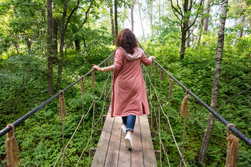 Young woman walking on suspension bridge in the forest. Traveler on the rope bridge. Travel woman on vacation concept. Hiking, summer outdoors activity