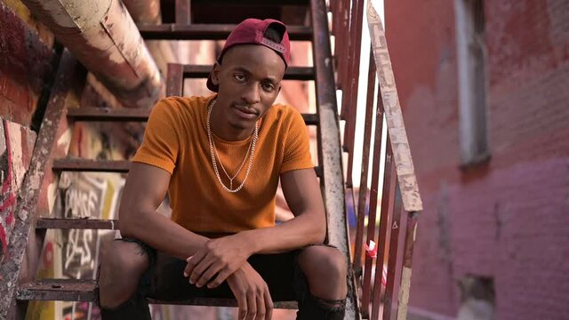 The camera zooms in on a young black guy sitting on a staircase in an alley in a dysfunctional neighborhood. Ghetto and young man. Slowmotion
