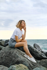 Fototapeta na wymiar A young attractive Caucasian blonde woman of 35 years sits on the rocks of sea coast. Emerald-colored water, waves splashing on the stones. The woman meditates and relaxes.