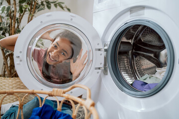 Smiling little girl with dark eyes peeks through round glass door of washing machine watches mom who is sorting laundry in bathroom fooling around playing having fun while doing household chores