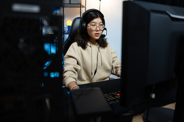 Focused gamer girl sitting in room in front of computer monitor professional gaming equipment headphones with microphone to talk to rivals team struggle to win strategy dark room with led lights