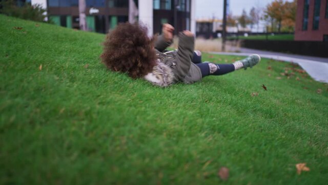 Little Afrcian American girl rolling down grassy hill outdoors in park in city in autumn day.