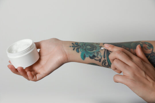 Woman applying cream on her arm with tattoos against light background, closeup