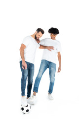 full length view of cheerful multiethnic men in jeans and t-shirts playing football on white.