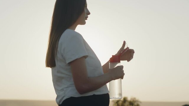 Pregnant woman stands in open air against background of sun and sky and drinks water from bottle, side view. Concept of proper nutrition