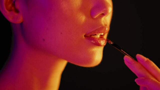Young Female Illuminated By Neon Light Applying Lip Gloss With Makeup Brush
