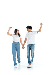 excited multiethnic couple showing win gesture while holding hands and walking on white.
