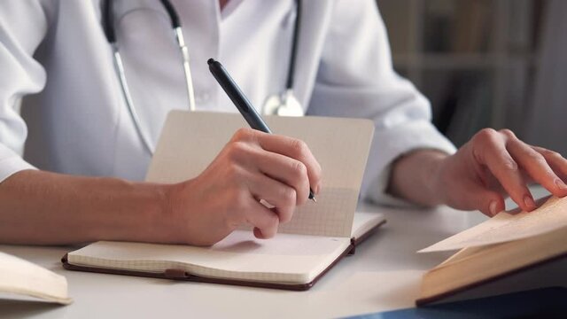 Medical research. Female doctor. Study education. Unrecognizable woman writing information in notebook from encyclopedia sitting light room interior.