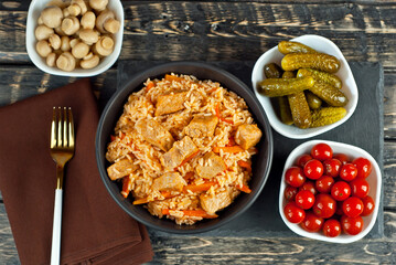 Pilaf with meat and carrots in a black bowl. Marinated mushrooms, tomato and cucumber on a black background. Food on a shabby table. Bowl on an old wooden board.