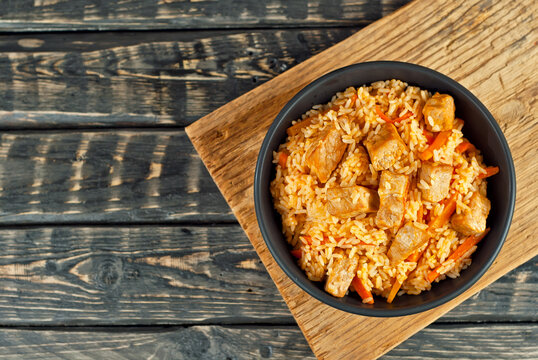 Pilaf with meat and carrots in a black bowl. Pilaf on a black background. Food on a shabby table. Bowl on an old wooden board. Copy space and free space for text near food.
