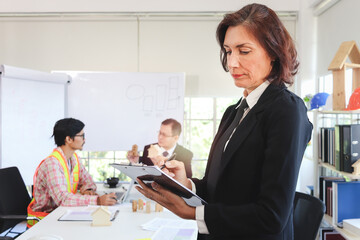 Confident beautiful middle aged business woman in suit holding document clip holder, writing file paper while standing in front of blurred background of business people brainstorming meeting in office