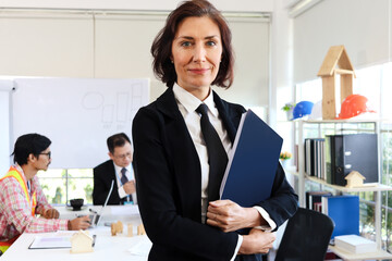 Confident beautiful middle aged business woman in black suit holding document folder and crossing arms while standing in front of business people brainstorming meeting blurred background in office